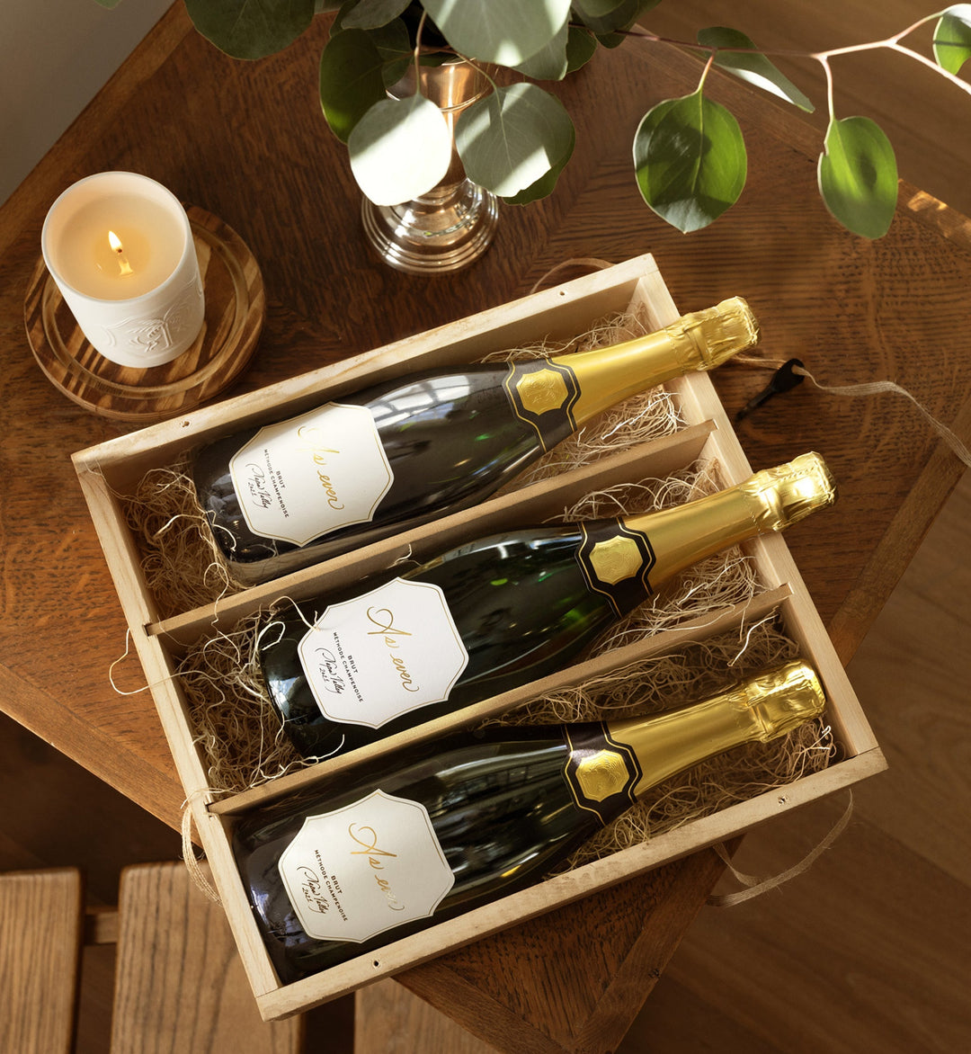 Three bottles of As ever wine in an As ever wooden gift crate on a wooden table with a candle and plant